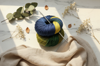 skein of yarn in blue and green with knitting needles on a light surface with leaves and flowers.